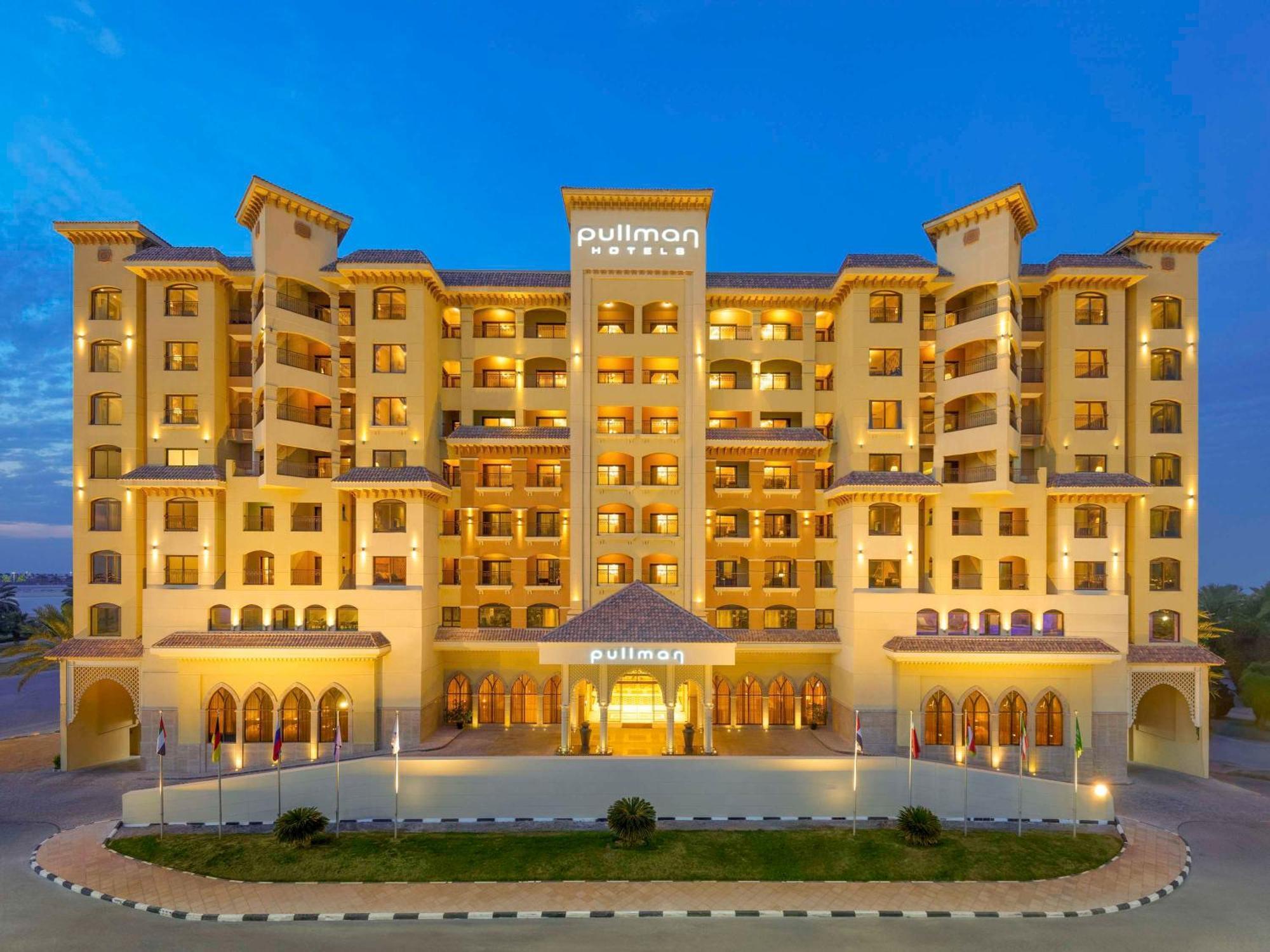 Pullman Resort Al Marjan Island Ras al-Khaimah Exterior photo The photo shows a modern hotel building, likely part of the Pullman brand. The structure features a warm, inviting color scheme with multiple floors and architectural details such as balconies and large windows. It is illuminated, creating a welcomin