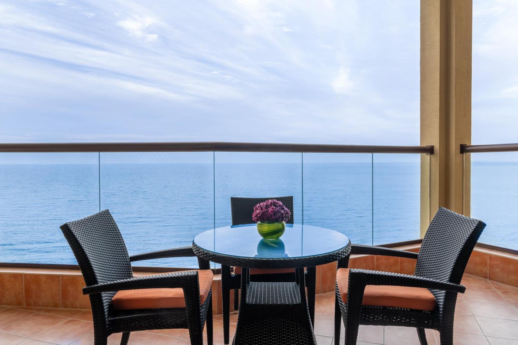 Pullman Resort Al Marjan Island Ras al-Khaimah Exterior photo The photo shows a small balcony or patio with a view of the ocean. There is a round glass table in the center with a green vase containing flowers. Two chairs with dark wicker frames and orange cushions are placed around the table. The backdrop featu