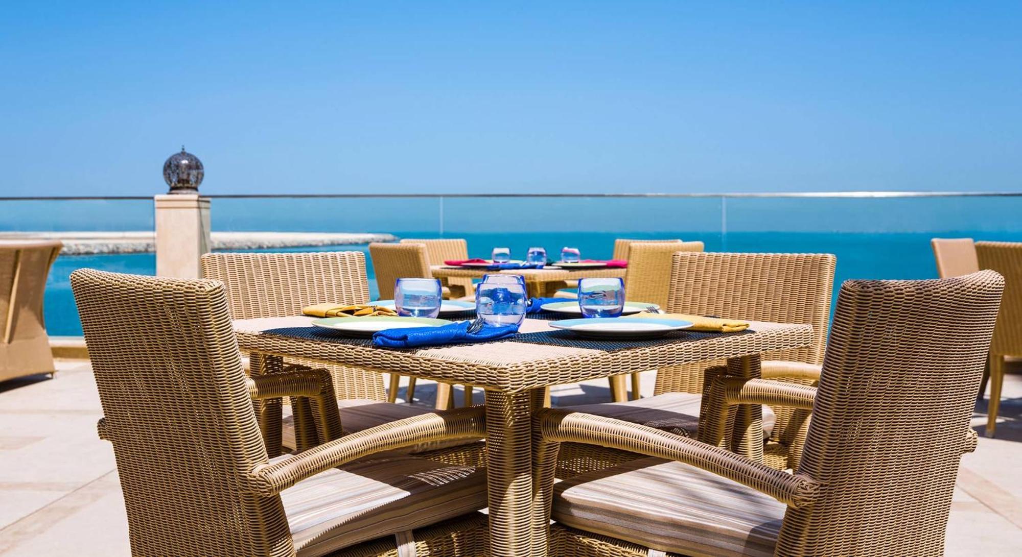 Pullman Resort Al Marjan Island Ras al-Khaimah Exterior photo The photo shows a set of outdoor dining furniture arranged on a patio or balcony with a view of the ocean. There is a square table with wicker chairs surrounding it. Each place setting includes a blue napkin, glasses, and dinner plates in vibrant col