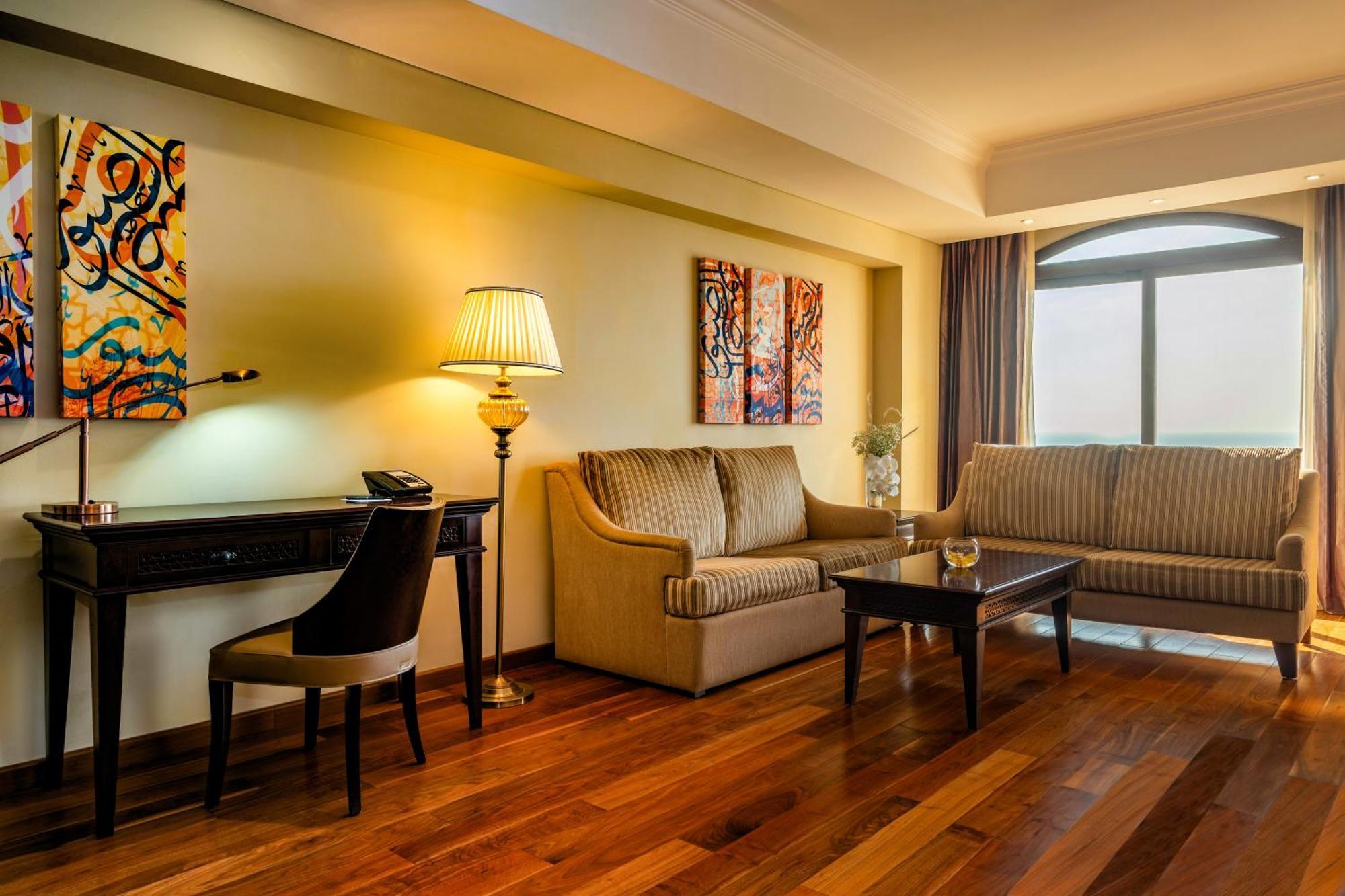 Pullman Resort Al Marjan Island Ras al-Khaimah Exterior photo The photo shows a cozy and stylish living area. It features two seating areas consisting of a light-colored sofa and a striped couch. There's a coffee table in the center made of dark wood. A desk with a lamp and a phone is positioned near the sofa. 