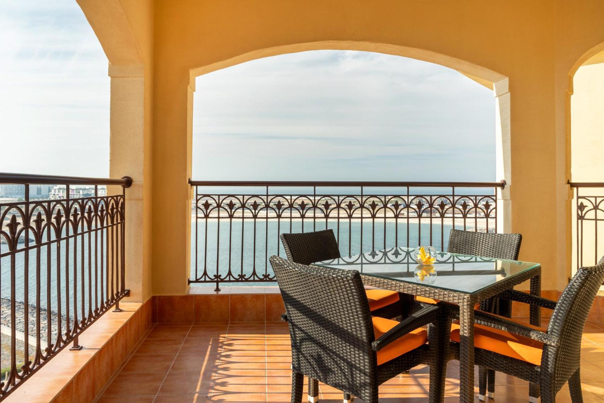 Pullman Resort Al Marjan Island Ras al-Khaimah Exterior photo The photo depicts a balcony or terrace area that features a glass-top table surrounded by four black wicker chairs with orange cushions. The space has an elegant wrought iron railing that overlooks a scenic view of the ocean or sea, visible through t