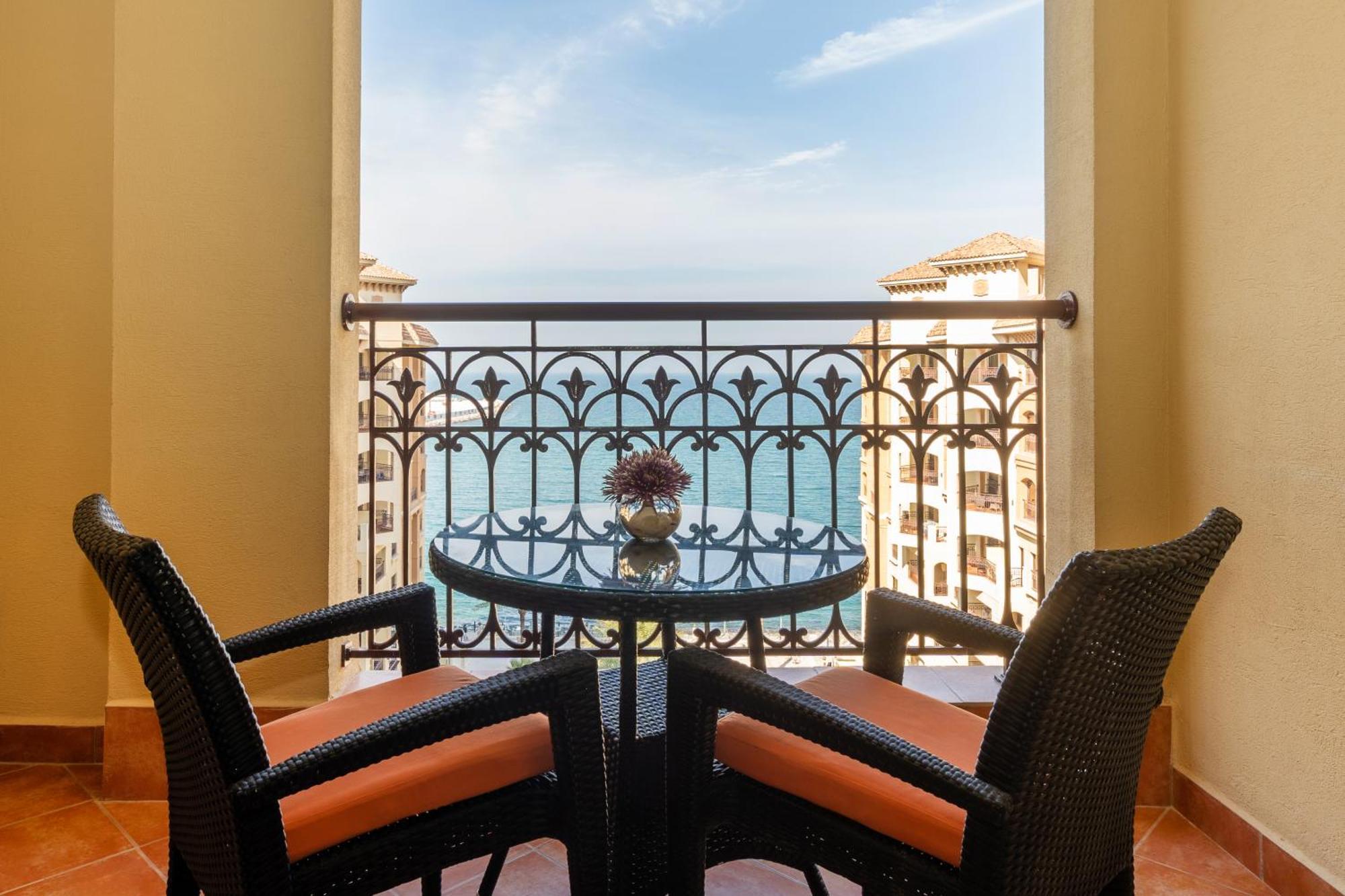Pullman Resort Al Marjan Island Ras al-Khaimah Exterior photo The photo shows a balcony scene featuring a small round table with two chairs. The chairs are made of wicker and have orange cushions. On the table, there is a small vase with a flower. In the background, there is a view of the ocean and a clear blue