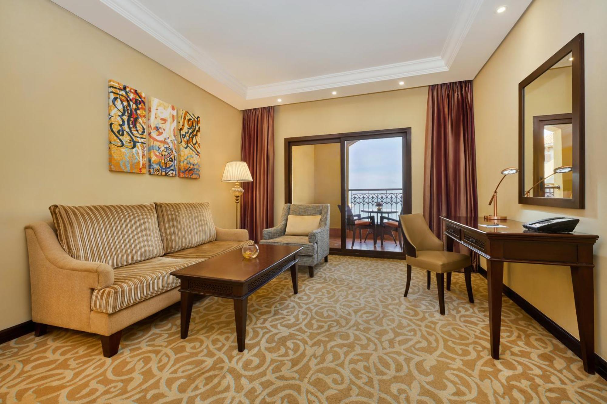 Pullman Resort Al Marjan Island Ras al-Khaimah Exterior photo The photo shows a cozy hotel suite. It features a seating area with a striped beige sofa and a gray armchair, arranged around a wooden coffee table. There's a lamp next to the sofa, and some colorful artwork is displayed on the wall. A mirror reflect
