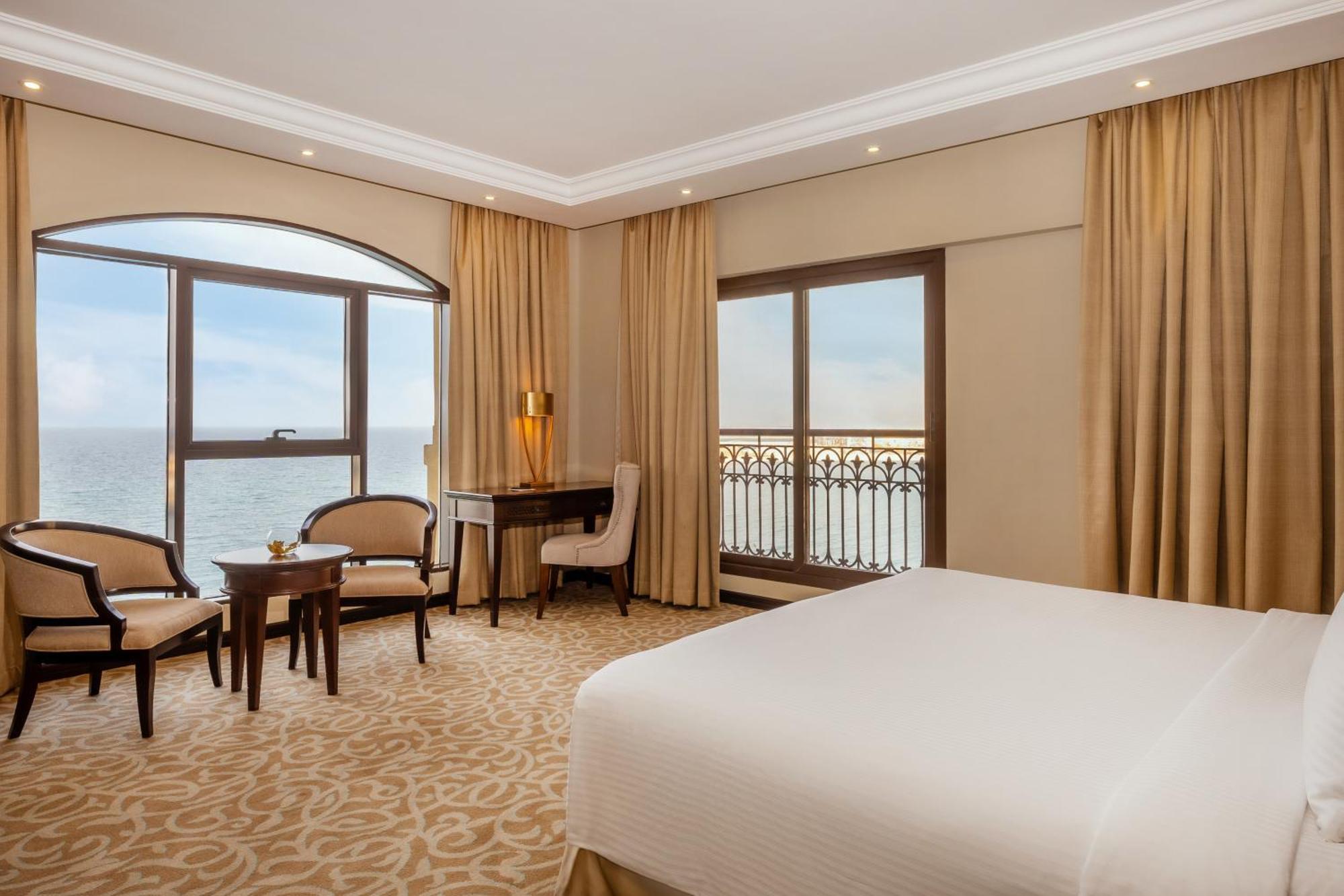 Pullman Resort Al Marjan Island Ras al-Khaimah Exterior photo The photo shows a hotel room with a modern and elegant design. It features a large bed covered with white bed linens, a seating area with two chairs and a small table near a window offering a view of the water outside. The room has light-colored wall