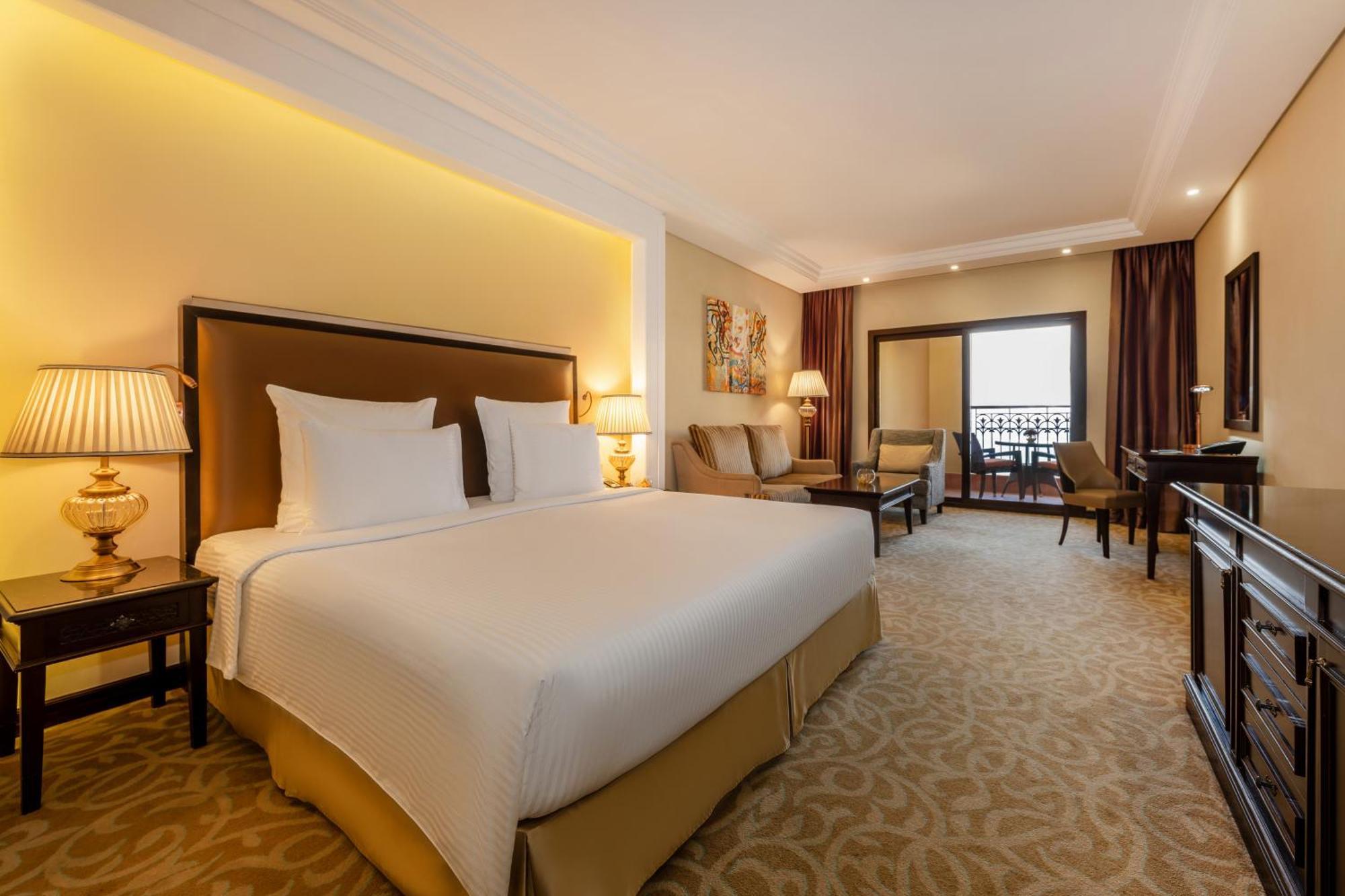 Pullman Resort Al Marjan Island Ras al-Khaimah Exterior photo The photo shows a hotel room featuring a large bed with white linens and a decorative headboard. The room is warmly lit, with soft yellow lighting accentuating the walls. There are two elegant bedside lamps on tables next to the bed. In the backgroun