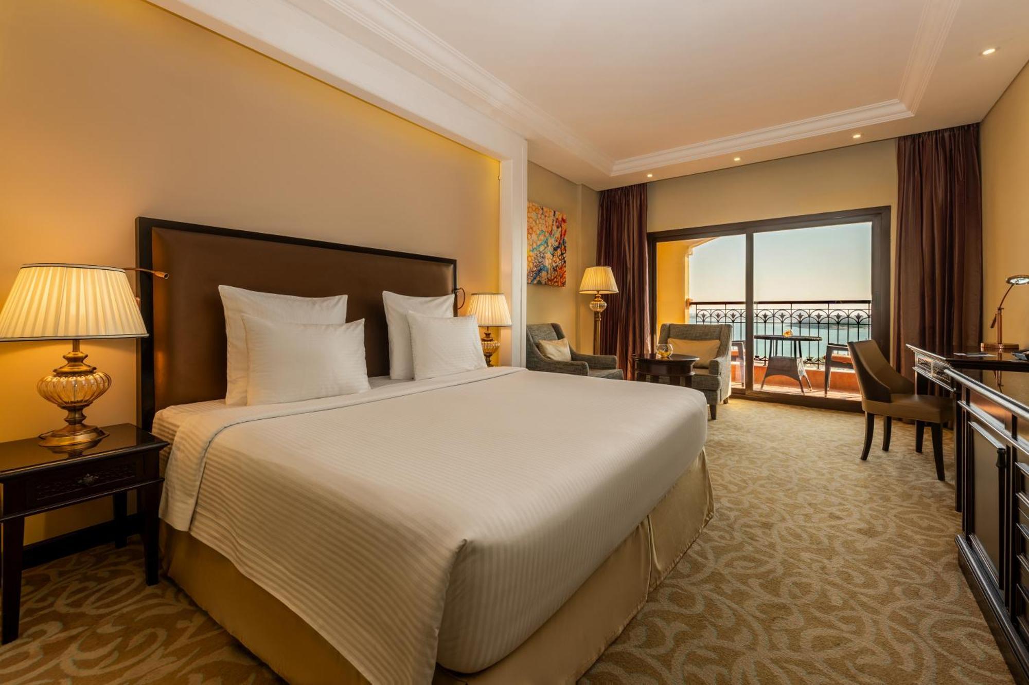Pullman Resort Al Marjan Island Ras al-Khaimah Exterior photo The photo shows a well-decorated hotel room. It features a large bed with crisp white bedding, two bedside lamps on either side, and a comfortable seating area with chairs. There are large windows that allow natural light to fill the space and provid