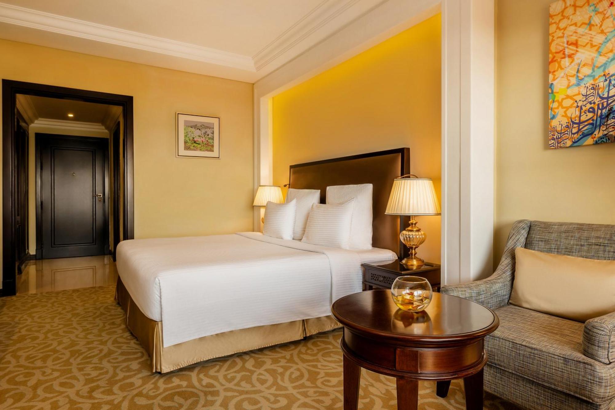 Pullman Resort Al Marjan Island Ras al-Khaimah Exterior photo The photo shows a neatly arranged hotel room featuring a double bed with white bedding, complemented by two bedside lamps with warm light. There's a small round table in front of the bed, likely with a decorative glass object on it. To the side, ther