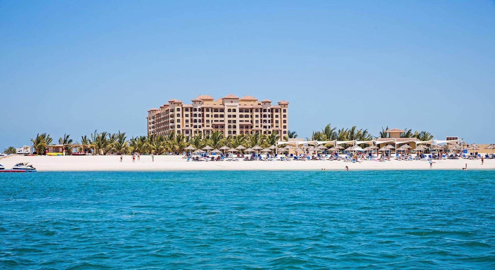Pullman Resort Al Marjan Island Ras al-Khaimah Exterior photo The photo shows a picturesque beach scene with clear turquoise water in the foreground. In the background, there is a large hotel or resort building with multiple stories, set against a clear blue sky. The beach features several umbrellas and lounge 