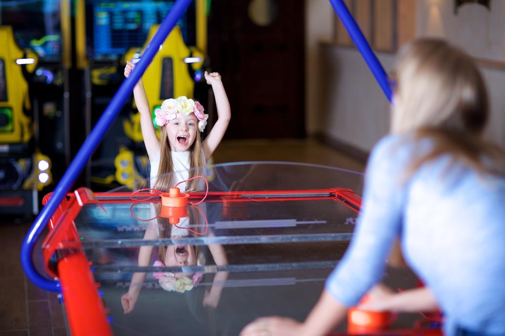 Pullman Resort Al Marjan Island Ras al-Khaimah Exterior photo The photo shows a joyful young girl with her arms raised in excitement, celebrating a win in an air hockey game. She is wearing a colorful flower crown and has a big smile on her face. Across from her, an adult woman is facing her at the air hockey t