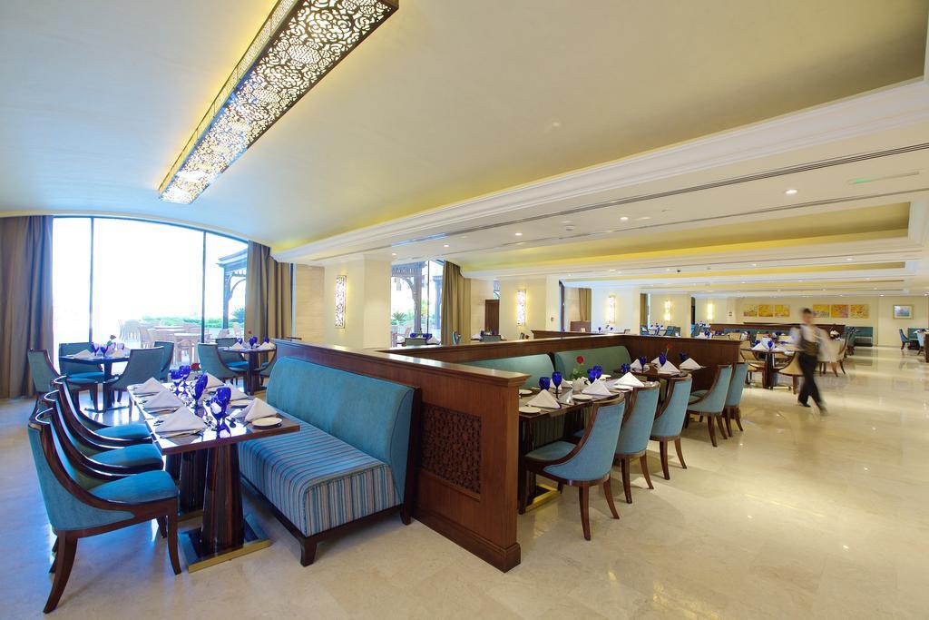 Pullman Resort Al Marjan Island Ras al-Khaimah Exterior photo The photo shows the interior of a restaurant. It features a spacious layout with a modern design. There are several dining tables arranged with blue glasses and neatly folded napkins. The seating includes a mix of upholstered chairs and a long, cushi