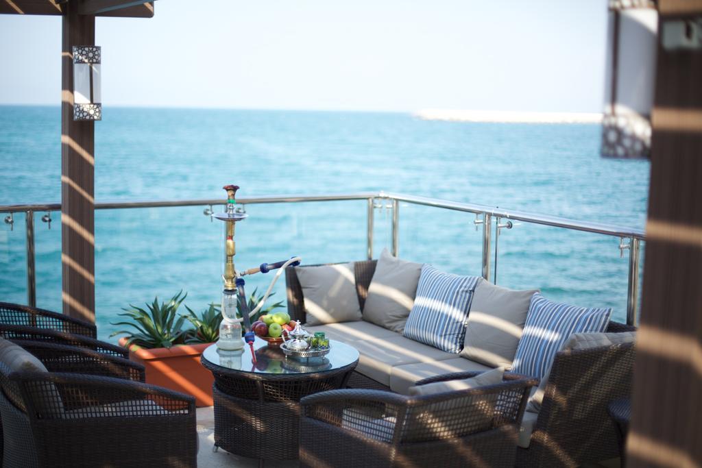 Pullman Resort Al Marjan Island Ras al-Khaimah Exterior photo The photo depicts a serene outdoor balcony or patio overlooking a body of water. In the foreground, there is a comfortable seating area with a light-colored sofa adorned with striped cushions. Surrounding the sofa are a few chairs, also designed for 