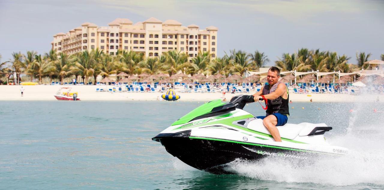 Pullman Resort Al Marjan Island Ras al-Khaimah Exterior photo The photo shows a man riding a jet ski on the water. He appears to be enjoying the activity, wearing swim shorts and holding onto the handles of the jet ski. In the background, there is a resort or hotel building with multiple stories, surrounded by 