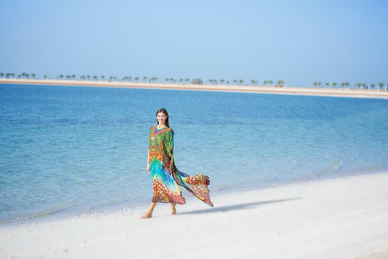 Pullman Resort Al Marjan Island Ras al-Khaimah Exterior photo The photo features a woman walking along a pristine, sandy beach with clear blue water in the background. She is wearing a colorful, flowing outfit that showcases vibrant patterns. The scenery demonstrates a picturesque coastal setting, with a bright