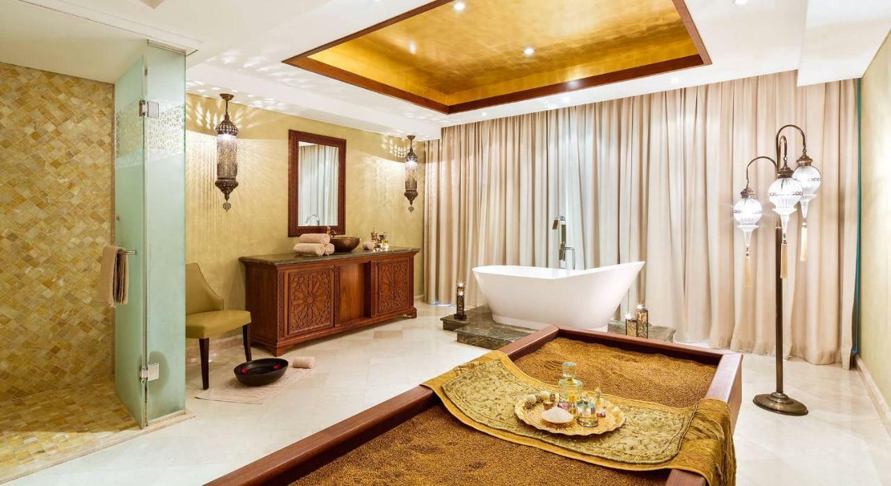 Pullman Resort Al Marjan Island Ras al-Khaimah Exterior photo The photo depicts a luxurious spa-like bathroom. It features a large, freestanding bathtub in the center, complemented by elegant gold accents on the ceiling. Along one side, there's a wooden vanity with a mirror, while light, flowing curtains provid